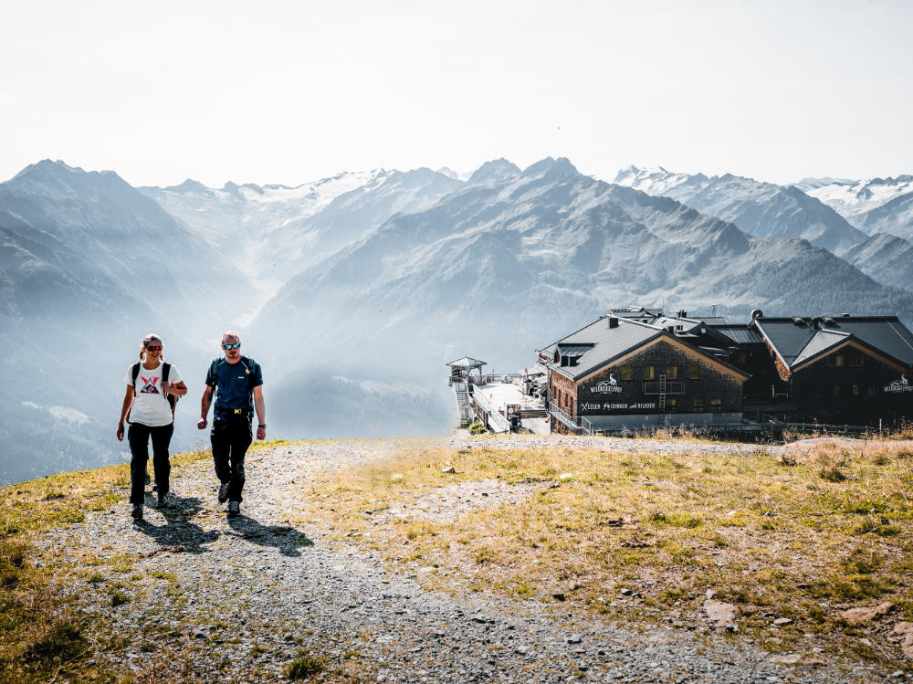 Vor der Kulisse der majestätischen Kitzbüheler Alpen verlassen zwei Wanderer eine Hütte am Wildkogel und begeben sich auf einen Pfad.