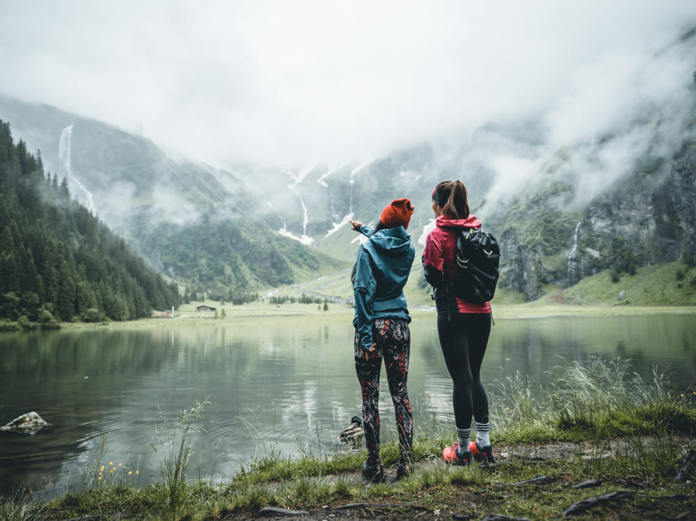 In der malerischen Landschaft von Mittersill stehen zwei Wanderinnen vor einem See, umgeben von Bergen und einem Wasserfall.