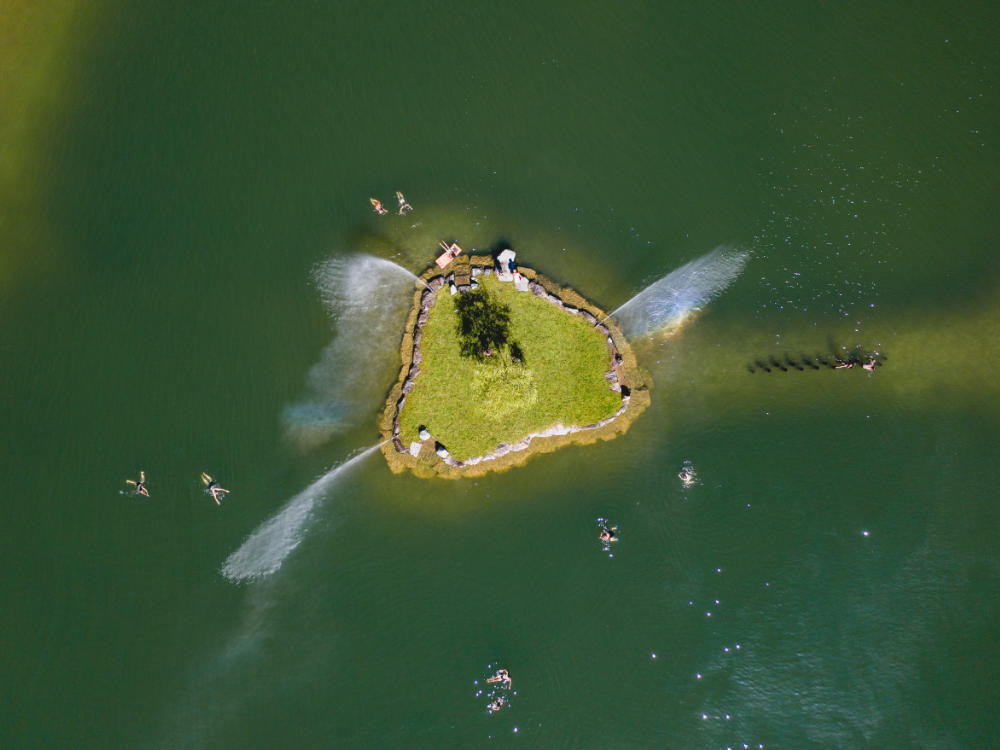 Luftaufnahme des Badesees Hollersbach mit einer kleinen Insel in der Mitte, umgeben von der malerischen Landschaft nahe Mittersill.