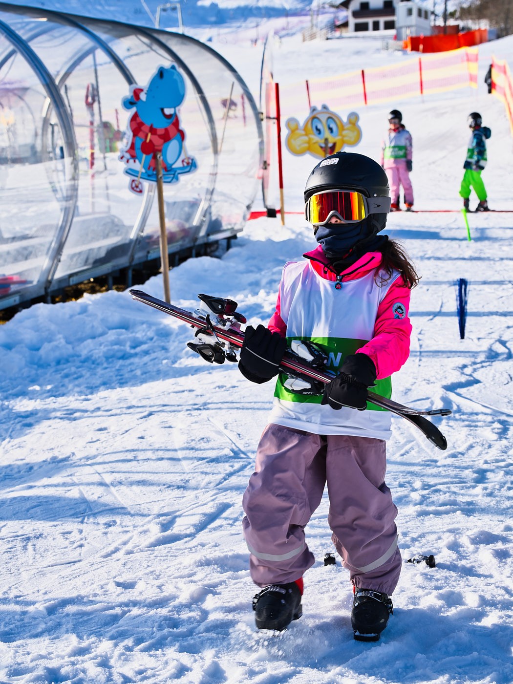 Kind mit Skihelm und Skibrille trägt Leihski am Übungshang der Skischule neben dem Zauberteppich