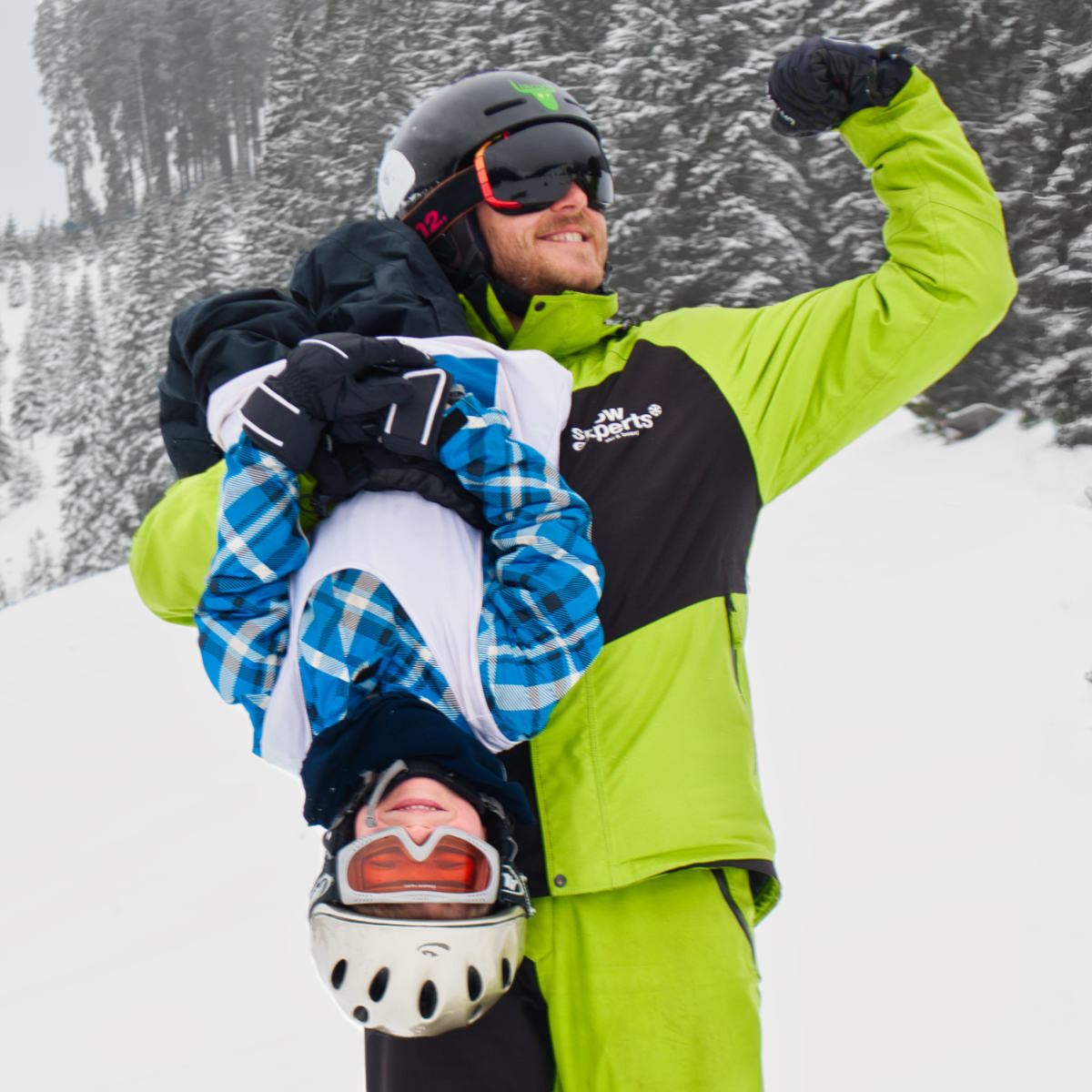 Snow Experts skileraar houdt een skicursuskind ondersteboven, beiden lachen en hebben duidelijk plezier in het KitzSki skigebied.